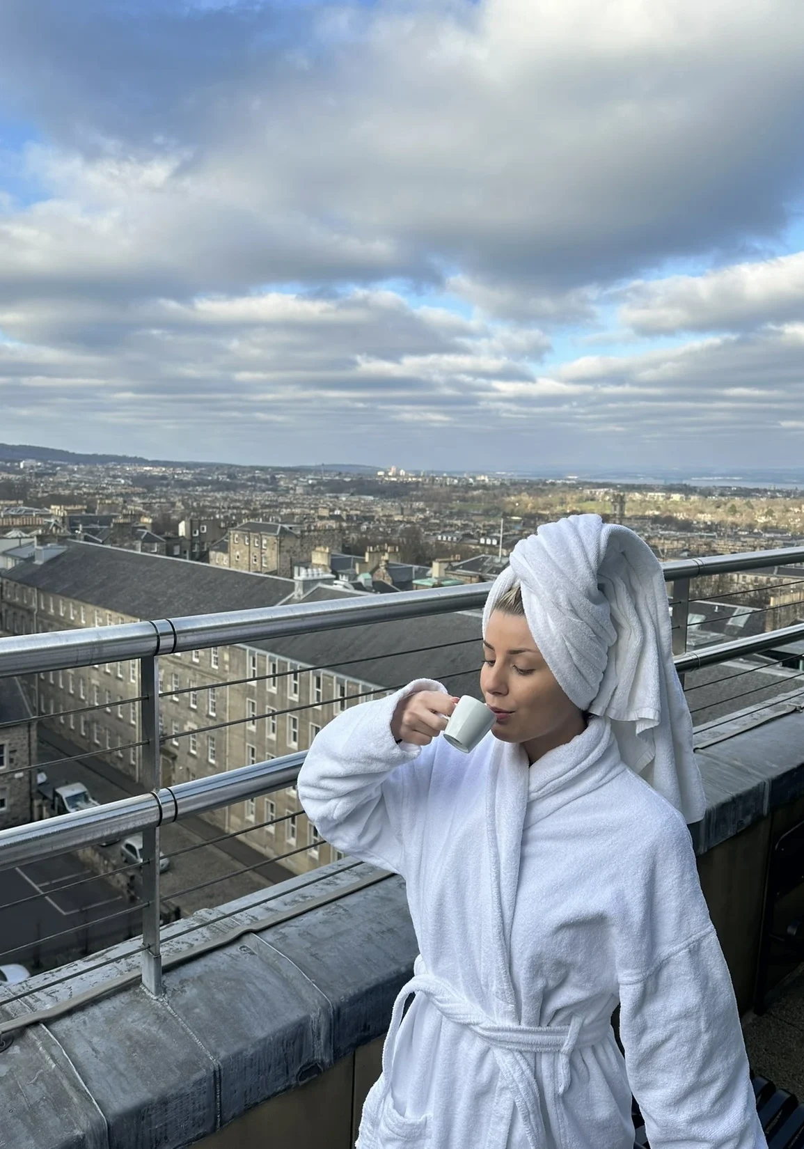 Woman in a white bathrobe drinking coffee on a balcony with panoramic city views in the background