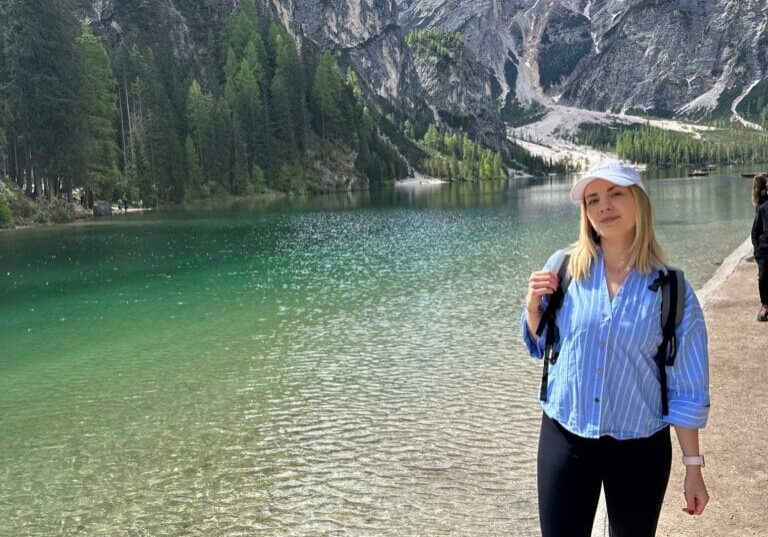 Hiking at Lago di Braies – Mountain Essentials Moment Woman hiking by Lago di Braies in the Dolomites wearing a backpack and cap, standing at the edge of a clear alpine lake.
