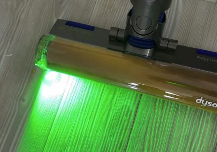 Dyson V12 Laser Slim Fluffy Head in Action – Dust Detection Technology Close-up of a Dyson V12 vacuum cleaner head with green laser light illuminating dust and debris on a light wood floor.